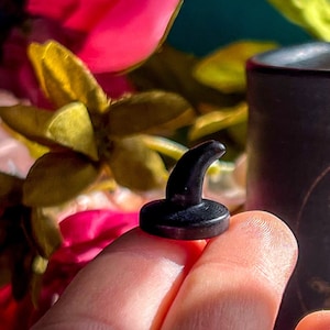 May include: A small black ceramic witch hat with a pointed tip, held in a hand against a background of pink and yellow flowers and a black mug with a gold moon design.