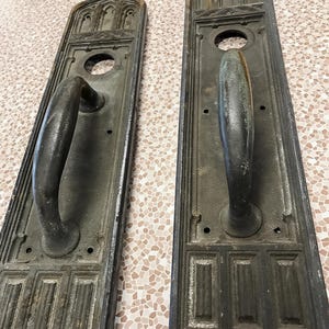May include: Two antique door handle plates with a dark, weathered finish. Each plate features an arched top with decorative details, a circular opening, and a curved handle. The handles are a darker shade than the plates.