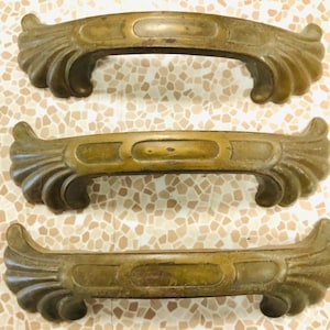 May include: Three antique brass cabinet door pulls with a decorative, ornate design. The pulls have a curved shape with a raised oval in the center and a scalloped edge.