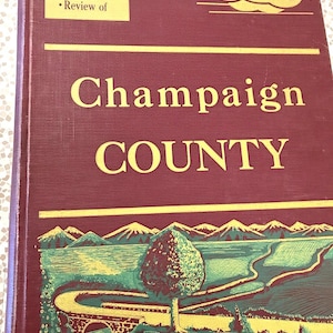 May include: A hardcover book titled Informative Historical Pictorial Review of Champaign COUNTY. The maroon cover features gold text and a green and yellow illustration of a landscape with mountains, a tree, a bridge, and a winding road.
