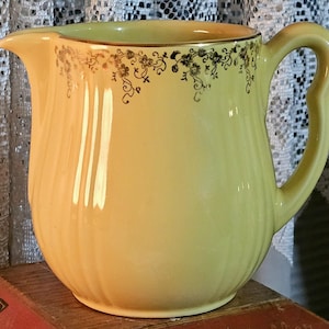 May include: A pale yellow ceramic pitcher with a curved handle and a spout. The pitcher has a gold rim and is decorated with a floral pattern in black and gold. The pitcher is sitting on a wooden surface.