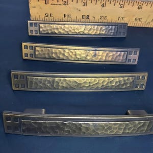 May include: Four bronze-toned cabinet pulls with a hammered texture. Each pull has a rectangular center and square accents on the ends. The pulls vary in size, with the longest measuring approximately 6 inches.