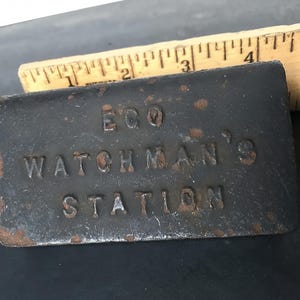 May include: A rectangular, dark gray metal object with the embossed text "ECQ WATCHMAN'S STATION." The object has a weathered appearance with rust-colored spots. A wooden ruler with inch markings is partially visible in the background.