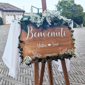 Può includere: Un cartello rettangolare in legno su un cavalletto in legno con il testo bianco Benvenuti, Mattia e Sara con un'icona a forma di cuore, e 9 Giugno 2023. Il cartello è decorato con fiori bianchi di nebbiolina, edera verde e drappi bianchi.