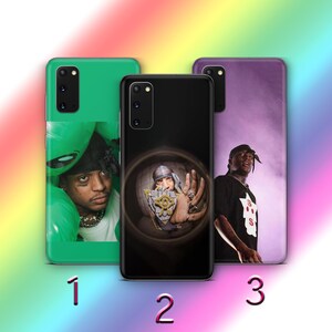 May include: Three different phone cases with different designs. The first case is green with a cartoon alien head. The second case is black with a photo of a person wearing a hat and sunglasses. The third case is purple with a photo of a person wearing a black shirt and a black hat.