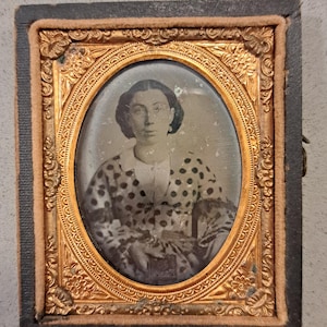 May include: Antique oval portrait photograph of a person in a polka dot dress, set within a decorative gold-toned frame. The frame is encased in a black rectangular case with a clasp. The photograph is a vintage piece, likely from the 19th century.