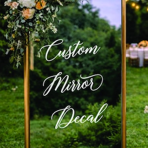 May include: A large rectangular mirror with a gold frame, adorned with a floral arrangement at the top. The mirror features white script that reads "Custom Mirror Decal FOR WEDDINGS & EVENTS". The background is a green lawn with trees.