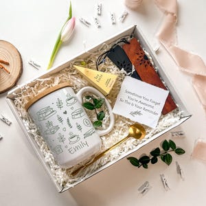 Puede incluir: Una taza blanca con tapa de bambú, con una ilustración verde y blanca de libros, tazas de café y hojas. La taza está personalizada con el nombre "Emily". La taza forma parte de una caja de regalo que incluye una bolsita de té, una cuchara, un marcapáginas y una nota que dice "Sometimes You Forget You're Awesome. So This Is Your Reminder."