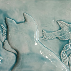 Könnte beinhalten: Eine Keramikfliese mit blauer Glasur, die einen Delfin zeigt, der durch Seegras schwimmt. Der Delfin befindet sich im Vordergrund und das Seegras im Hintergrund. Die Fliese ist hellblau und hat eine glänzende Oberfläche.