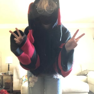 May include: A person wearing a black and red striped hoodie with a black face mask and clear glasses. The hoodie has large, floppy ears. The person is making a peace sign with both hands.