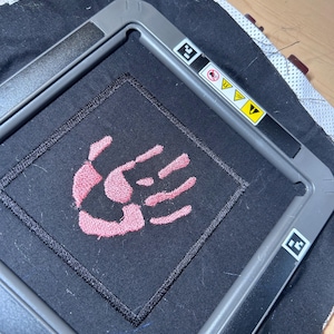 May include: A pink hand-shaped embroidery design on a black fabric background. The design is in a square frame.