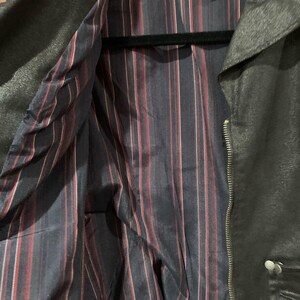 May include: Black jacket with a silver zip and button fastening. The interior lining features navy blue fabric with red and white vertical stripes. The jacket is hanging on a black hanger.