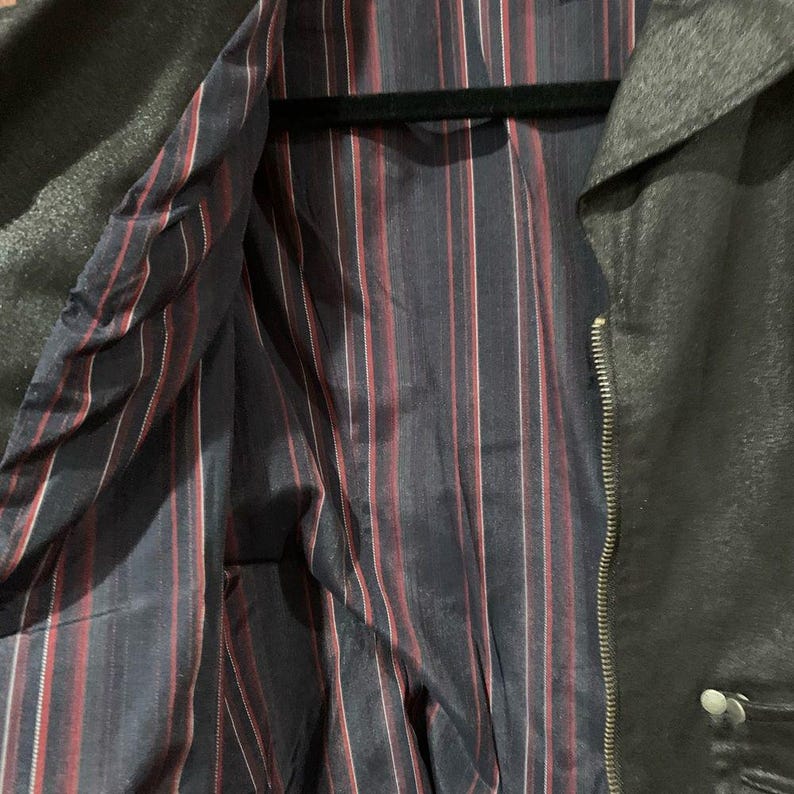 May include: Black jacket with a silver zip and button fastening. The interior lining features navy blue fabric with red and white vertical stripes. The jacket is hanging on a black hanger.
