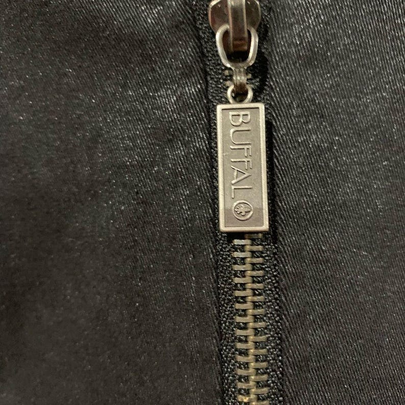 May include: Close-up of a black garment zipper with a silver metal pull. The pull tab is rectangular and features the word "BUFFALO" in capital letters. The zipper teeth are a matching silver colour.
