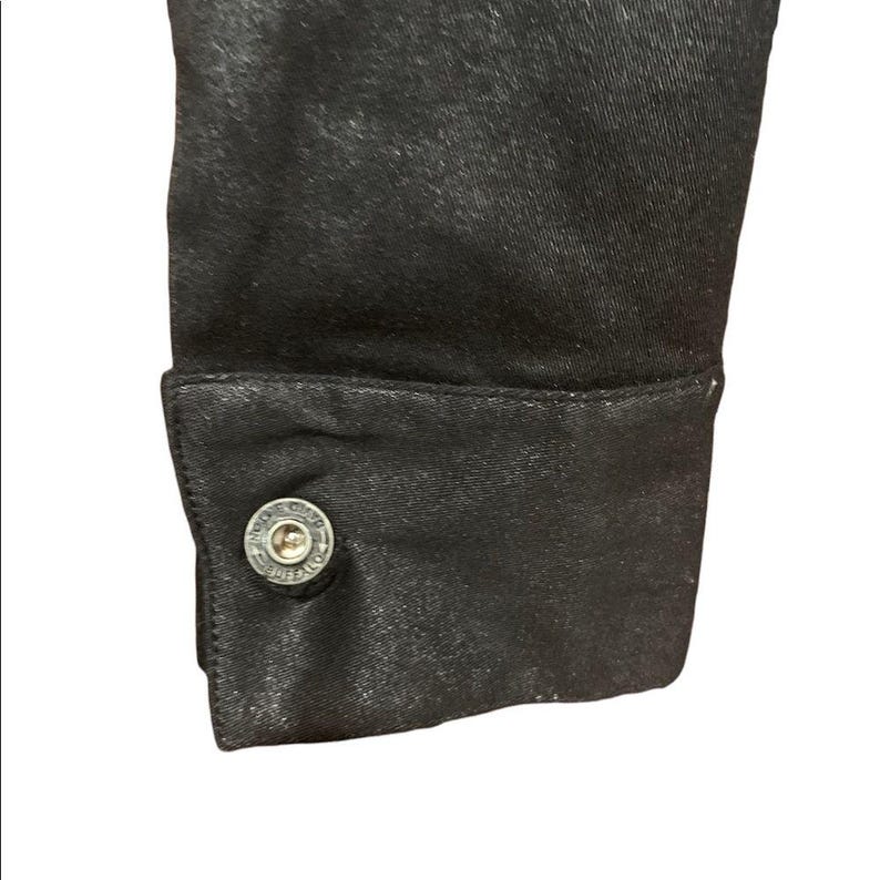 May include: Detail of a black garment sleeve and buttoned cuff. The cuff has a silver button with embossed text. The fabric has a slight sheen, possibly satin or a similar material.