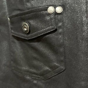 May include: Close-up of a black garment with a pocket and two silver buttons. The pocket has a single silver button. The fabric has a subtle sheen, and the stitching is visible. The buttons are round with a decorative design.