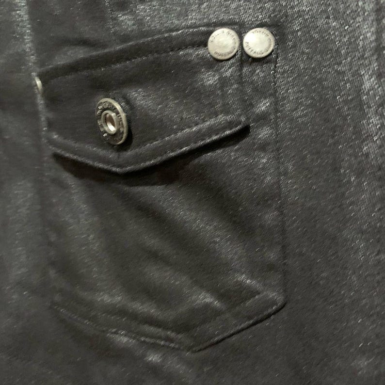 May include: Close-up of a black garment with a pocket and two silver buttons. The pocket has a single silver button. The fabric has a subtle sheen, and the stitching is visible. The buttons are round with a decorative design.