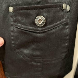 May include: Close-up of a black jacket pocket with a silver button that says "DAVIDSON" and two smaller silver buttons. The pocket is rectangular and has a flap. The jacket appears to be made of a shiny material.