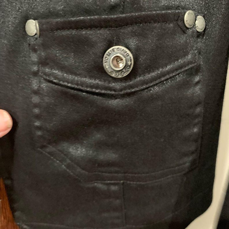 May include: Close-up of a black jacket pocket with a silver button that says "DAVIDSON" and two smaller silver buttons. The pocket is rectangular and has a flap. The jacket appears to be made of a shiny material.