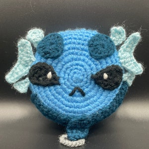 May include: A blue crocheted plush toy with a sad expression. The toy has large black eyes, a small frown, and two light blue crocheted fins on the sides.