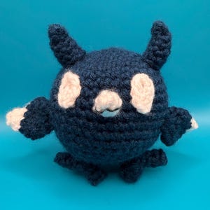 Puede incluir: Una criatura de ganchillo hecha a mano en azul marino con detalles blancos. El cuerpo redondo tiene dos parches blancos en forma de ojo, brazos pequeños y dos pies pequeños. La criatura tiene dos cuernos pequeños y una boca blanca. El fondo es azul claro liso.