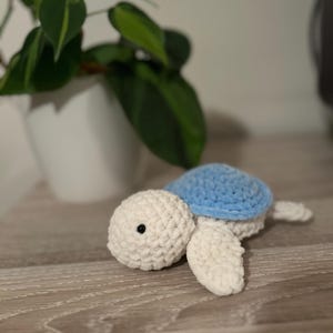 Op de afbeelding: Een gehaakt knuffeldier schildpad met een wit lichaam en een blauwe schild. De schildpad ligt op een houten oppervlak.