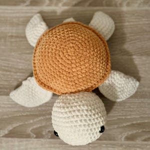 Op de afbeelding: Een gehaakt schildpad speelgoed met een wit hoofd en een oranje schild. Het speelgoed is gemaakt van zacht garen en heeft zwarte ogen.