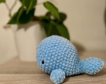 May include: A light blue crocheted whale plush toy with black eyes. The whale is lying on a wooden surface.