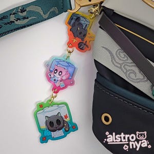 Op de afbeelding: Een kleurrijke sleutelhanger met drie bedels met cartoonkatten. De bedels zijn bevestigd aan een turquoise en witte band. De bedels zijn gemaakt van helder acryl met een regenboogverloop. De eerste bedel toont een zwarte kat met een witte buik en een roze neus. De tweede bedel toont een roze kat met een witte buik en een blauwe neus. De derde bedel toont een zwarte kat met een witte buik en een gele neus. De bedels houden allemaal verschillende voorwerpen vast, zoals een camera, een boek en een rol toiletpapier. De sleutelhanger is bevestigd aan een zwarte tas met een gouden rits en een gouden metalen ring.