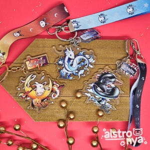 May include: Four colorful keychains with different designs. Each keychain features a clear acrylic charm with a white rabbit, a blue dragon, a black snake, and a red and white Chinese character. The keychains are hanging on a wooden surface with gold beads scattered around.