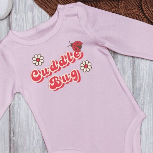 Puede incluir: Mono de bebé rosa de manga larga con el texto "Cuddle Bug" en una fuente retro con una mariquita y flores.