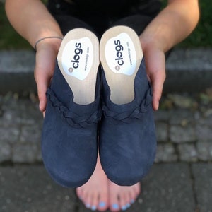 Könnte beinhalten: Ein Paar marineblaue Clogs mit einem geflochtenen Riemen über dem Vorderfuß und einem hellen Holzfußbett. Auf der weißen Einlegesohle ist der Text "clogs" und "mkclogs.com" aufgedruckt. Das Schuhwerk hat eine matte Oberfläche und abgerundete Zehen.