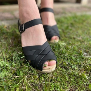 Może przedstawiać: Czarne skórzane sandały typu clog z odkrytymi palcami i zapięciem na klamrę. Sandały są noszone na trawie.