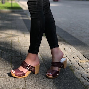 Könnte beinhalten: Ein Paar braune Ledersandalen mit einem Holzabsatz und einem Schnallen-Detail. Die Sandalen werden von einer Person getragen, die eine schwarze Jeans trägt.