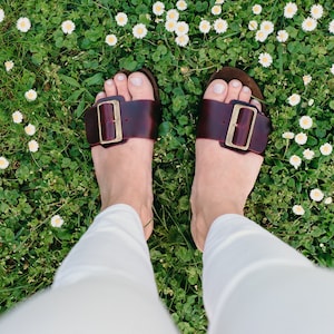 May include: A pair of brown leather sandals with gold buckles. The sandals are worn by a person standing in a field of white daisies.
