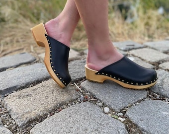 Puede incluir: Zuecos de cuero negro con suelas de madera y tachuelas doradas. Los zuecos se llevan puestos en los pies de una persona, de pie en un camino empedrado.