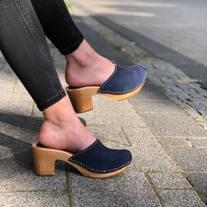 May include: Navy blue suede clogs with wooden heels. The clogs have a rounded toe and are accented with gold-colored studs along the edge of the sole. The heels are approximately 3 inches high. The clogs are worn with black jeans.