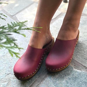 May include: Burgundy clogs with wooden soles and brass-coloured stud accents. The clogs have a rounded toe and a slip-on design. The image shows the clogs on a stone surface.