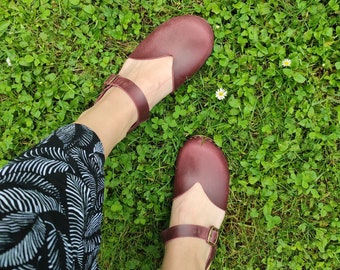 May include: A pair of dark burgundy leather clogs featuring a rounded toe, a deep V-shaped cutout on the vamp, and an adjustable ankle strap with a metallic buckle. The clogs have a smooth finish and are worn with black and white patterned trousers.