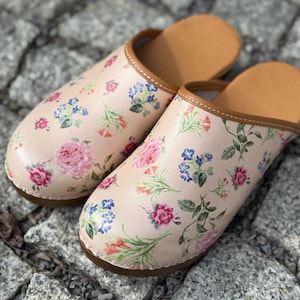 Puede incluir: Un par de zapatillas con estampado floral. Las zapatillas tienen un color base rosa claro con un patrón repetido de rosas rosas, flores azules y naranjas y hojas verdes. El interior y el ribete son marrones.