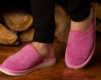 May include: A pair of bright pink suede slippers featuring a perforated design on the upper, a rounded toe, and a flat, textured white rubber sole. The slippers have a matching pink trim around the opening and a soft interior lining.