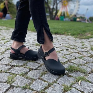 May include: Black leather clogs with a strap and buckle closure. The clogs have a rounded toe and a small heel. The shoes are worn with black pants. The background shows a park with a Ferris wheel.