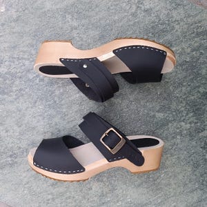 May include: Black leather clogs with a wooden platform and two straps. The straps have a buckle closure. The clogs have a peep toe.