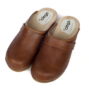 Può includere: Un paio di zoccoli in pelle marrone con suola in legno e un cinturino sulla parte superiore. Gli zoccoli hanno un dettaglio di cucitura bianca attorno alla suola e un'etichetta bianca all'interno dello zoccolo che dice "clogs" e "www.myclogs.com".