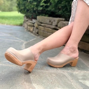 May include: Beige leather clogs with wooden heels. The clogs have a closed toe and slip-on design. The heels are approximately 7.6 cm tall. The clogs are displayed outdoors on a stone surface.