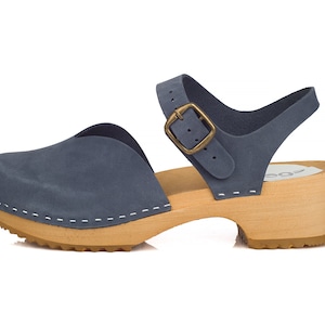 May include: Blue suede clog sandal with a wooden sole and a strap with a brass buckle. The sandal has a closed toe design and a small heel. The sole is made of light-colored wood, and the upper is a muted blue.