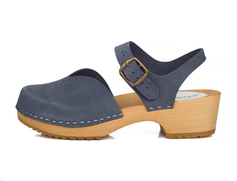 May include: Blue suede clog sandal with a wooden sole and a strap with a brass buckle. The sandal has a closed toe design and a small heel. The sole is made of light-colored wood, and the upper is a muted blue.