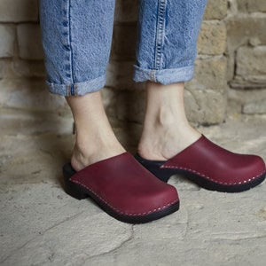 May include: A pair of burgundy leather clogs with a wooden sole. The clogs are worn by a person standing on a concrete floor.