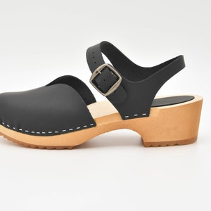 May include: Black leather clog sandal with a wooden heel and a buckle closure. The sandal features a closed toe design, an ankle strap, and a light brown sole. The heel height is approximately 2 inches.