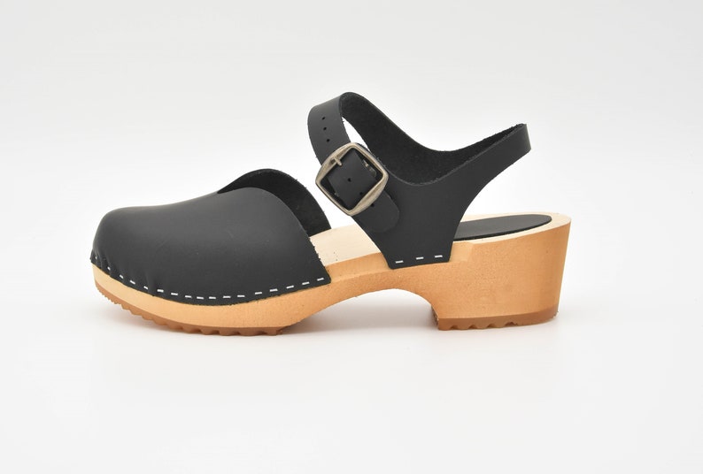 May include: Black leather clog sandal with a wooden heel and a buckle closure. The sandal features a closed toe design, an ankle strap, and a light brown sole. The heel height is approximately 2 inches.